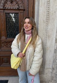 Veste matelassée beige clair, écharpes rose et marron, jeans bleu, sac à bandoulière jaune avec sangle assortie, debout près d'une porte en bois ornée avec des détails.