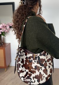 Barna-fehér foltos válltáska ívelt formával, bőr pánttal, arany színű f�ém részekkel és puha tapintású anyagból, zöld pulóveren viselve.