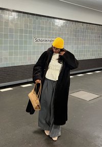 Person wearing a yellow knit hat, glasses, black coat, white top, and grey trousers, carrying a beige handbag, standing at a subway station near the Schönleinstraße sign.