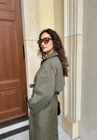 Ελαιόχρωμο καπιτονέ παλτό με κοντό σχέδιο, ζωνάκι στη μέση και φαρδύ γιακά. Συμπληρωμένο με στυλάτα καφέ γυαλιά ηλίου με σχέδιο χελώνας.