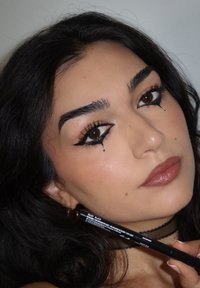 Kobieta z czarnym eyelinerem w stylu skrzydełek i małymi kroplami pod oczami trzyma czarny ołówek do eyelinera blisko twarzy.