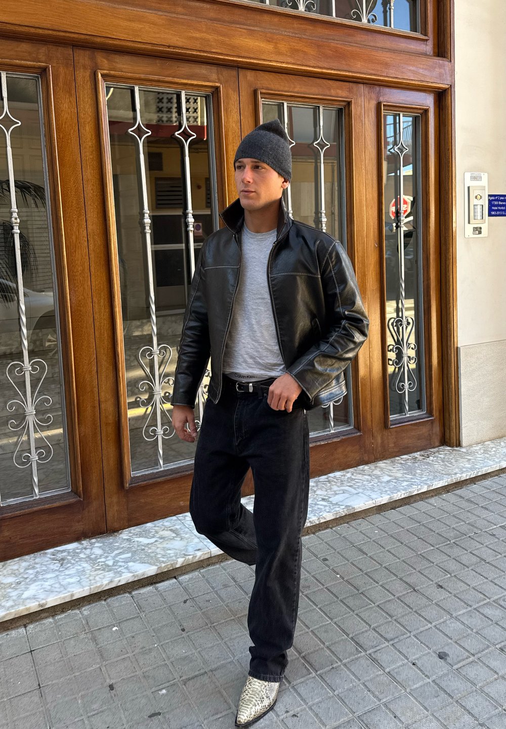 Férfi fekete bőrdzsekiben, szürke sapkában, szürke ingben, fekete farmerben és kígyóbőr mintás csizmában, amint városi járdán áll.