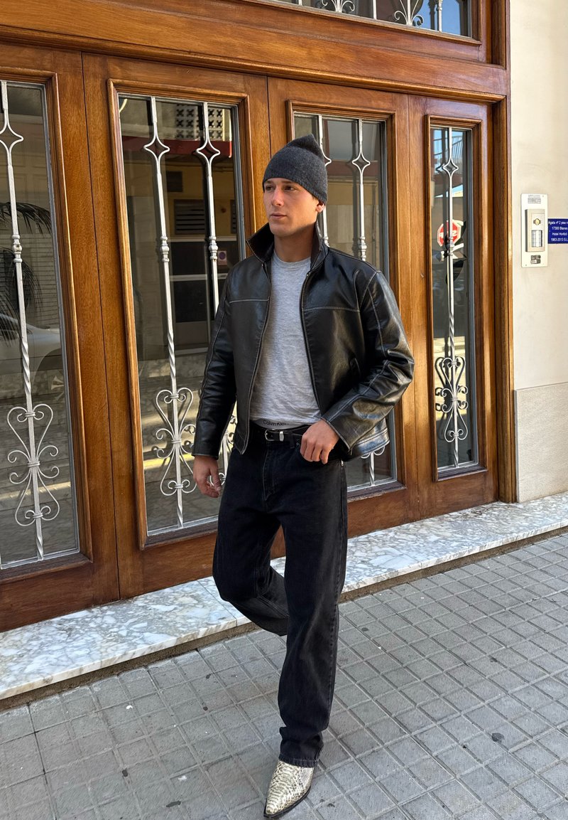 Man iklädd svart läderjacka, grå mössa, grå tröja, svarta jeans och stövlar med ormskinnsmönster står på stadens trottoar.