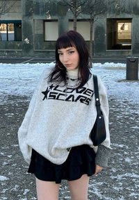 Pulôver cinza oversized com letras pretas "LOVE SCARS," emparelhado com uma saia preta em camadas. Os acessórios incluem colares em camadas e uma pequena bolsa preta.