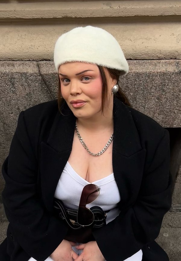 Junge Frau mit weißem Barett, schwarzem Mantel, weißem Oberteil, silberner Kettenkette und großen Ohrringen, die an einer Steinmauer sitzt.
