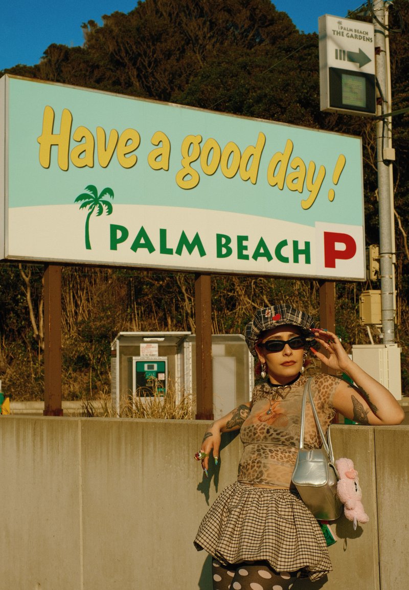 Personne avec tatouages et lunettes de soleil posant près d'un panneau « Have a good day! Palm Beach » dans un cadre extérieur avec des arbres et des équipements de stationnement.