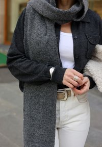 Grijze gebreide cardigan met bijpassende lange sjaal, wit tanktopje eronder en crème-kleurige broek. Opvallende zilveren ring aan de hand.