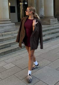 Brązowa, oversize'owa marynarka na burgundowej, dopasowanej sukience, zestawiona z szarymi trampkami i białymi skarpetkami. Stoi na kamiennych schodach w pobliżu kolumn.