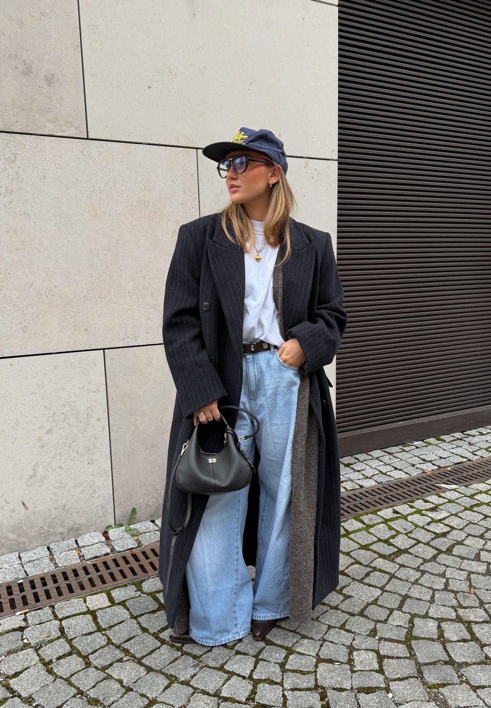 Long manteau noir à fines rayures porté sur un T-shirt blanc, jean bleu clair à jambes larges, sac à main noir et casquette bleu marine avec texte jaune. Lunettes de soleil carrées.
