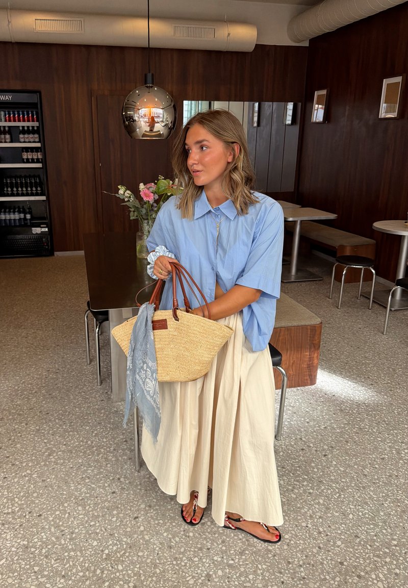 Femme en blouse bleue et jupe crème tenant un sac en paille avec une écharpe bleue, debout dans un café moderne avec des murs et des tables en bois.