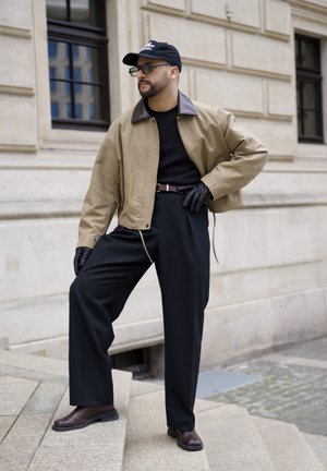 Hombre con chaqueta beige, camisa negra, pantalones anchos negros, guantes de cuero, gorra y gafas de sol posa en unos escalones de piedra junto a un edificio.