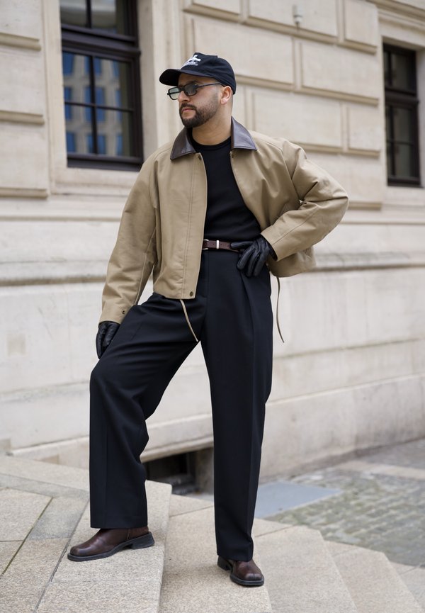 Mand iført beige jakke, sort skjorte, brede sorte bukser, læderhandsker, kasket og solbriller poser på stentrappen ved en bygning.