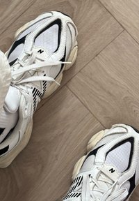 Λευκά sneakers με πολυεπίπεδη σχεδίαση, μαύρες λεπτομέρειες, πλέγματα και υφασμένες σόλες. Οι κορδόνες είναι χαλαρά δεμένες. Ξύλινο δάπεδο στο φόντο.