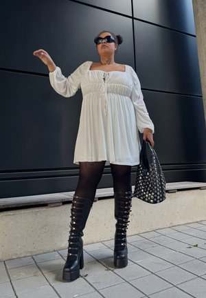 Vestido mini blanco, fluido, con mangas abullonadas y un lazo en el escote, combinado con botas de muslo negras texturizadas y un bolso con tachuelas.