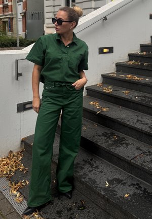 Camisa de algodón verde de manga corta con bolsillo, combinada con pantalones de pierna ancha de talle alto a juego. Zapatos negros de punta y gafas de sol.