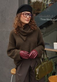 Pull marron à col bénitier, gants bordeaux, sac à main vert olive et b éret noir. Cheveux bouclés et lunettes complètent le look.