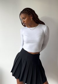 Weisses langärmliges Crop-Top, kombiniert mit einem schwarzen Faltenrock. Das Top ist glatt, während der Rock tiefe Falten und einen elastischen Bund hat.
