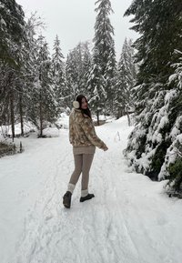 Faux-Fur-Jacke mit einem beige-braunen Muster, cremefarbenem Pulli, grauen Leggings und schwarzen Stiefeln, in einer verschneiten Waldlandschaft.