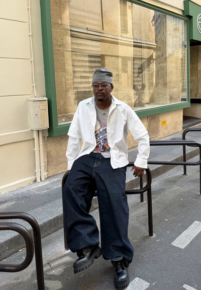 Camisa blanca de botones con rayas verticales sobre una camiseta gráfica, combinada con jeans de mezclilla oscuros de pierna ancha y botas de plataforma negras. Los accesorios incluyen gafas y un gorro gris.