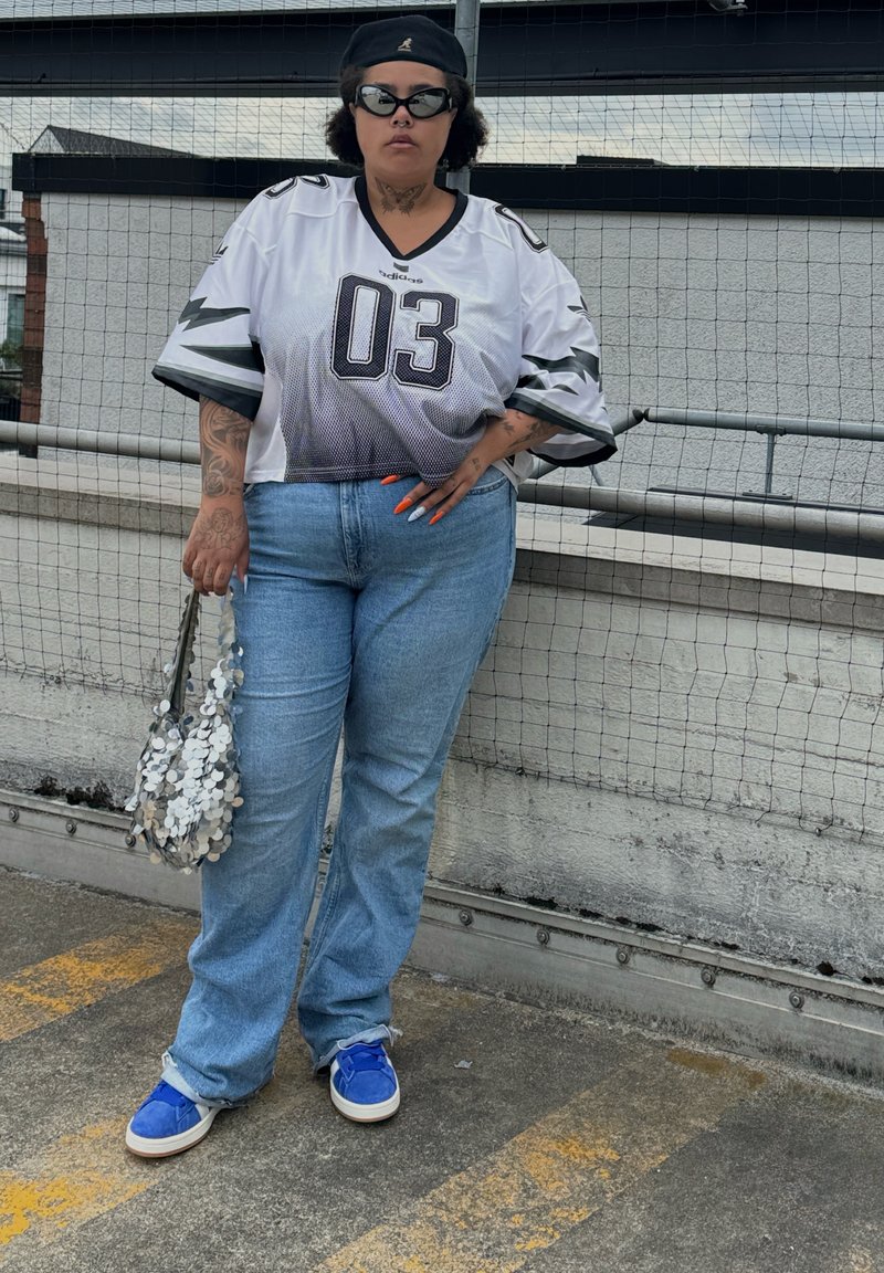 Camisa de futebol oversized branca com detalhes pretos e o número 03, jeans azul claro, sapatos azuis e uma bolsa prateada com lantejoulas.