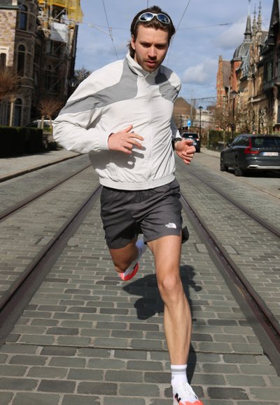 Hombre con chaqueta blanca y gris y pantalones cortos negros corriendo por una calle empedrada con vías de tranvía, edificios urbanos y coches estacionados al fondo.
