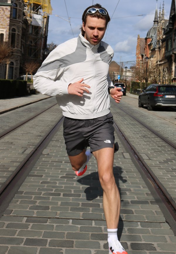 Mann in weißer und grauer Jacke und schwarzer Shorts, der auf einer Kopfsteinpflasterstraße mit Straßenbahnschienen läuft; im Hintergrund sind städtische Gebäude und geparkte Autos zu sehen.
