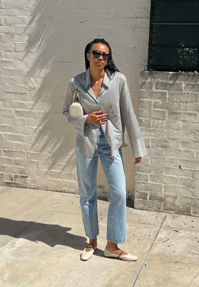 Mujer con camisa de rayas grande, vaqueros azul claro, zapatos planos beige, gafas de sol y bolso blanco pequeño de pie frente a una pared de ladrillos clara.