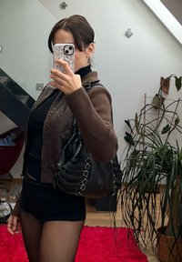 Καφέ δερμάτινο μπουφάν με ριγωτά μανίκια, μαύρη μπλούζα και μαύρη τσάντα ώμου με διακοσμητικές λεπτομέρειες στον ιμάντα. Καθρεφτισμένο φόντο με φυτά.
