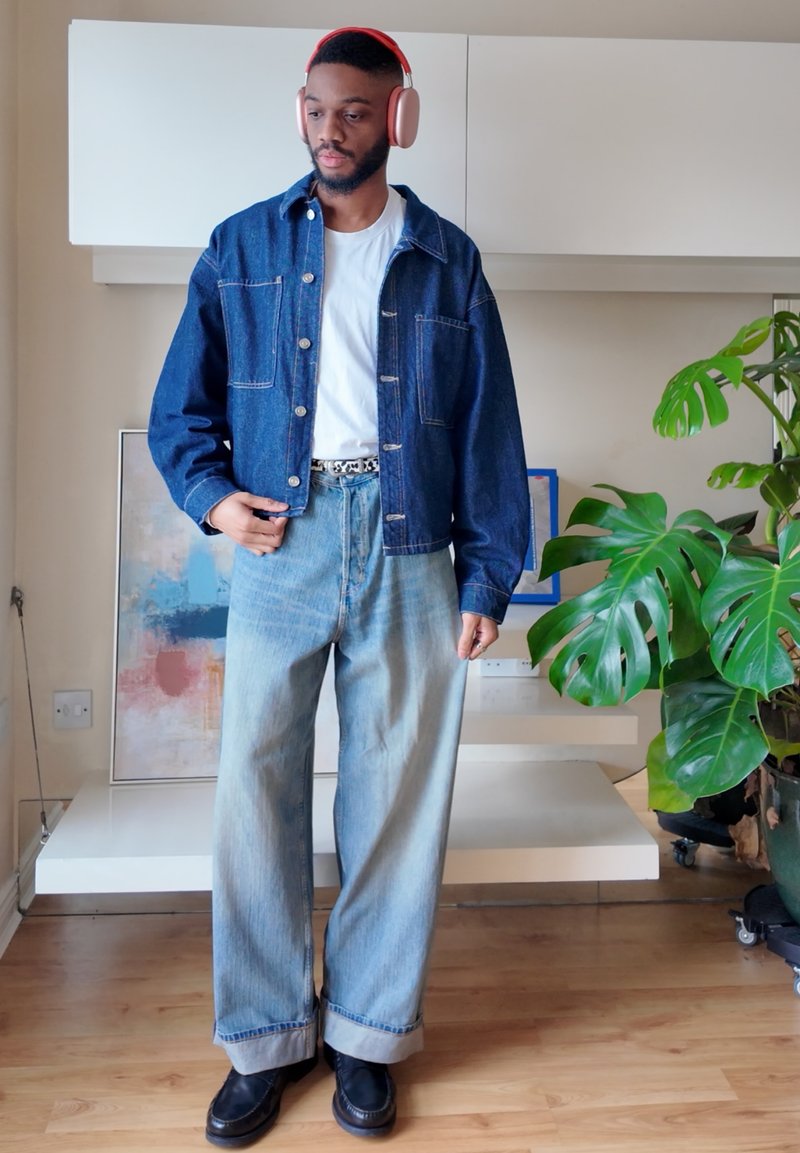 Homme portant un casque rouge, une veste en jean, un t-shirt blanc, un jean à jambes larges et des chaussures noires, debout près d'une grande plante verte à l'intérieur.