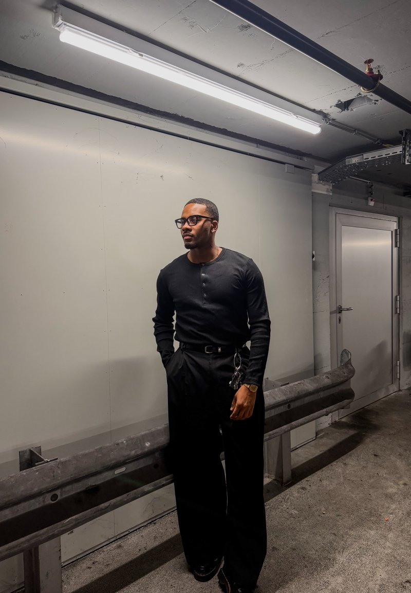 Chemise noire à manches longues côtelée, associée à un pantalon noir ample. Caractérisée par un col simple et un porte-clés accroché au pantalon. Fond neutre.