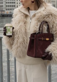 Sac à main en daim bordeaux avec poignées supérieures, accents en métal doré, tenu par une personne portant un pull en maille beige et un manteau duveteux, tenant une tasse de café.