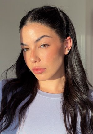 Retrato de una persona con cabello largo y oscuro, que lleva una camisa color púrpura claro, con labios ligeramente brillantes y múltiples piercings en las orejas.