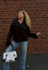 Καφέ κοντό μπουφάν με φαρδύ γιακά, συνδυασμένο με ανοιχτό γαλάζιο τζιν. Μία υφασμάτινη λευκή τσάντα με μαύρες κηλίδες ολοκληρώνει το σύνολο. Το φόντο είναι από τούβλα.