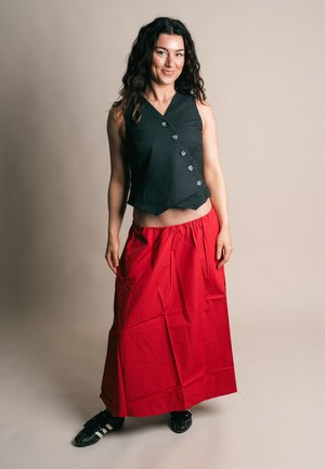 Mujer con cabello largo y rizado, que lleva una blusa negra sin mangas con botones, falda larga roja y zapatillas negras Adidas, de pie contra un fondo liso.