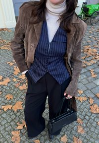 Brūns zamšādas mētelis pār tumsā zilu svītrainu vestu un krēmkrāsas augsto apkakli, kombinēts ar plašām melnām biksēm un melnu somiņu.