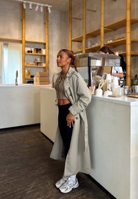 Lys beige trenchcoat over en strikket brun crop top og svarte tights. Skoene er sølv- og hvite sneakers. Moderne kafé-setting med treakenter.