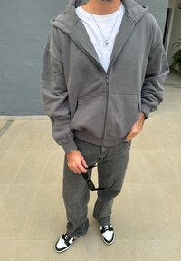 Moletom cinza claro com fecho, com grandes bolsos, usado sobre uma t-shirt branca. Combinado com jeans cinza escuro e ténis preto e branco. Óculos de sol na mão.