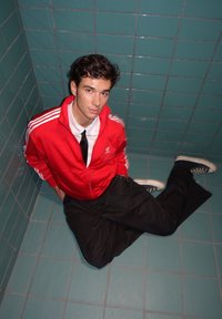 Rood sportjack met witte strepen, zwarte broek en sneakers. Zittend tegen een groene tegelmuur, met het haar nonchalant gestyled.