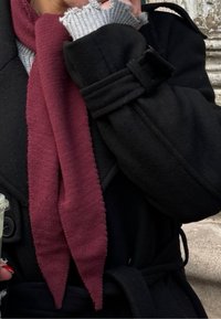 Personne en manteau noir ajustant une moufle grise avec la main gauche, portant une écharpe rouge foncé et tenant un gobelet en plastique contenant une boisson verte.