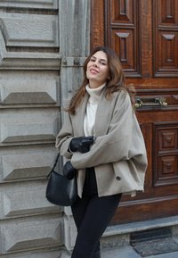 Beige oversized frakke med sjalskrave, sorte læderhandsker og en sort h�åndtaske. Baggrunden har tekstureret sten og en trædør.