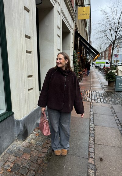 Abrigo de gran tamaño, de color marrón oscuro y textura, con botones, pantalones de pierna ancha grises y un bolso rosa. De pie en una calle de adoquines, sonriendo.