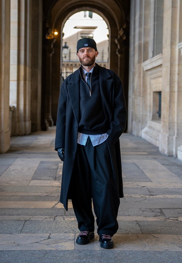 Homme portant un pardessus noir, un pantalon large, des gants et une casquette, marchant dans un couloir voûté en pierre, les mains dans les poches.