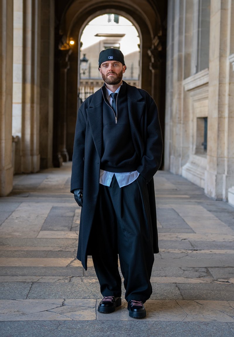Muž v čiernom kabáte, širokých nohaviciach, rukaviciach a súdčku kráča chodníkom so kamennými oblúkmi so založenými rukami v vreckách.