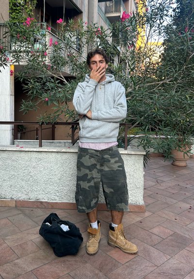 Sudadera con capucha gris, pantalones cortos de camuflaje y botas de trabajo marrones. El fondo presenta vegetación verde y flores rosas, con una pared de piedra texturizada.