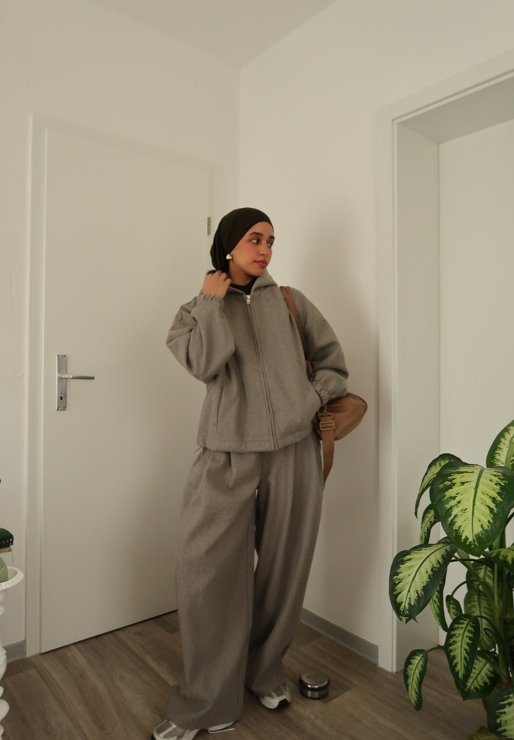 Veste polaire grise zippée et pantalon ample assorti, accompagnés d'une écharpe sombre et d'un sac à bandoulière beige. Cadre intérieur avec une plante à proximité.