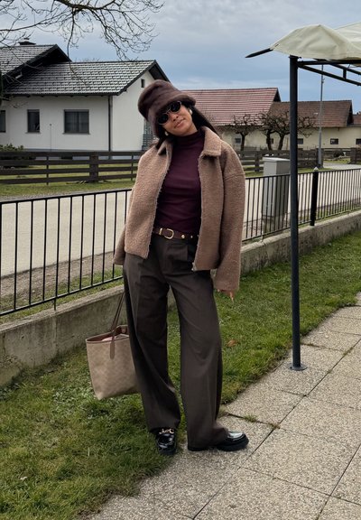 Mujer con chaqueta marrón, boina, pantalones oscuros y gafas de sol, de pie sobre el césped al lado de una calle residencial cercada bajo un cielo nublado.