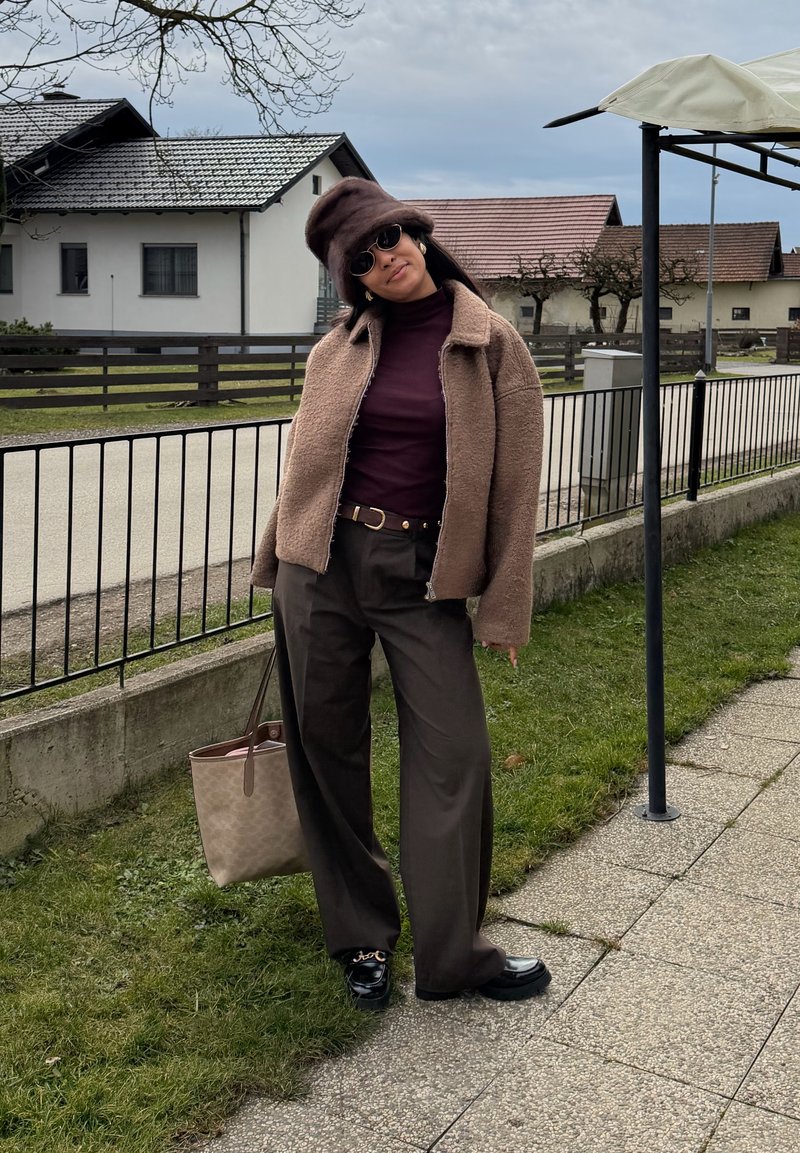 Femme portant une veste marron, un béret, un pantalon foncé et des lunettes de soleil, debout sur l’herbe à côté d’une rue résidentielle clôturée sous un ciel nuageux.