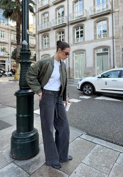 Chaqueta bomber verde oliva sobre una camiseta blanca, combinada con unos pantalones vaqueros holgados de color gris oscuro. Sosteniendo una taza, de pie contra un farola en una calle de la ciudad.