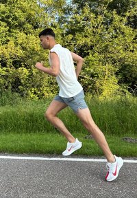 Valged sportlikud jalanõud punaste detailidega, kerge ilma varrukateta särk ja hallid lühikesed püksid, mida kannab jooksja rohelise muru äärel.