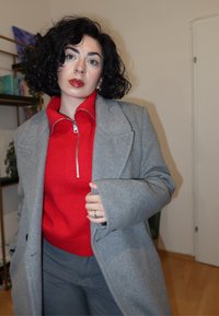 Szürke gyapjúkabát piros cipzáras pulóver fölött, szürke nadrággal párosítva. Göndör fekete haj, kezében a kabát hajtókáját tartva, semleges belső térben áll.